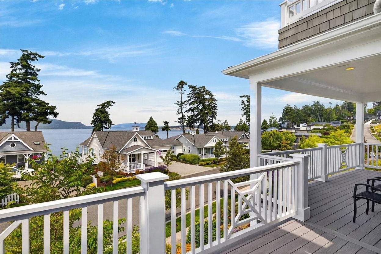 This image captures a scenic view from a covered balcony, showcasing a coastal neighborhood with charming houses nestled near the water. The balcony features a white railing and gray decking, providing a comfortable vantage point to overlook the lush landscaping and the distant bay. The bright, clear sky and expansive water view create a serene and inviting atmosphere typical of a premium waterfront property.