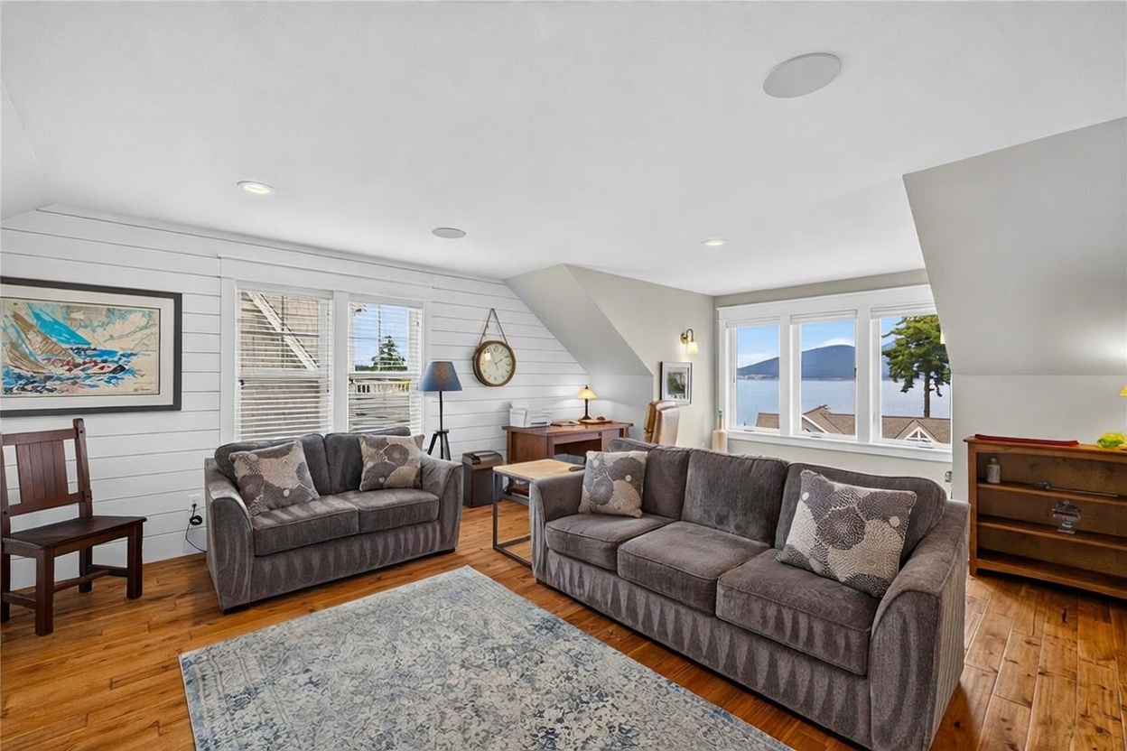 This bright and airy living room features white shiplap walls, warm wood flooring, and a large area rug, creating a cozy yet coastal-inspired atmosphere. The space is furnished with two plush grey sofas and a small wooden desk area, while large windows provide a stunning, unobstructed view of the water and distant mountains. The room is illuminated by recessed lighting and a stylish floor lamp, offering a comfortable and inviting space for relaxation.