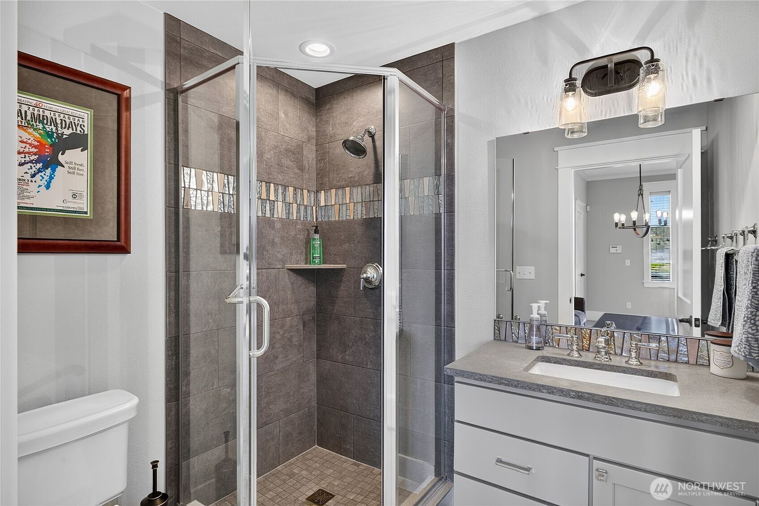 This bathroom features a corner shower enclosure with dark, textured wall tiles and a decorative mosaic accent strip. A vanity with a stone countertop and a rectangular undermount sink sits beneath a large mirror, which reflects the adjacent room. The space is illuminated by a dual-light fixture above the mirror and a recessed light over the shower, creating a clean and functional aesthetic.
