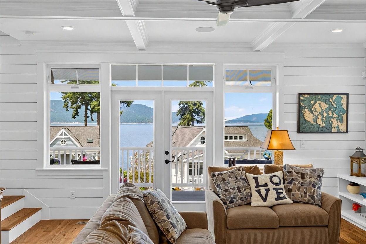 This bright living room features a comfortable brown sofa adorned with decorative pillows, including one with 'LOVE' lettering. The space is framed by large windows and a central glass-paned door that provide an expansive, unobstructed view of the water and distant mountains. White shiplap walls and a coffered ceiling enhance the coastal aesthetic, while a framed nautical map adds a thematic touch to the decor.