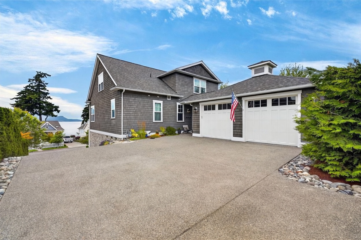This front-facing view captures a multi-level home with gray shingle siding and a prominent two-car garage featuring white doors and a cupola. The property is situated on a sloped lot with a concrete driveway leading to the entrance, while a scenic water view is visible in the background. The architectural style is classic coastal, complemented by manicured landscaping and a clear blue sky.