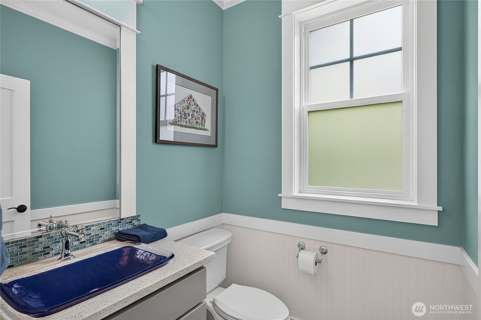 This compact powder room features a distinctive blue glass vessel sink set atop a light-colored stone countertop. The walls are painted a soft teal, complemented by white wainscoting and a framed piece of art, while a window with frosted glass provides natural light. The space is finished with a white toilet and a simple, clean aesthetic.