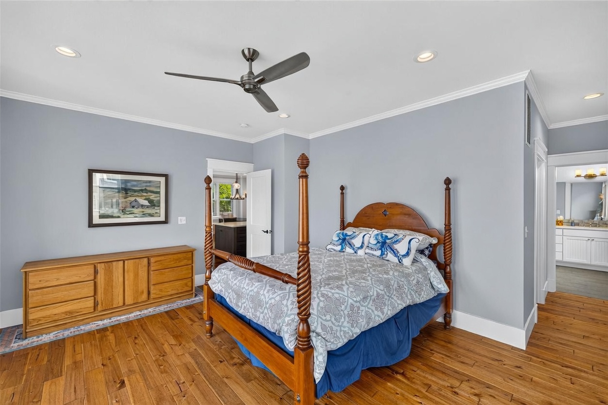 This primary bedroom features a classic four-poster wooden bed as the centerpiece, complemented by light blue walls and warm-toned wood flooring. A wooden dresser sits against the wall beneath a framed landscape painting, while a matching nightstand with a lamp provides bedside lighting. The room includes a ceiling fan and offers a glimpse into an adjacent bathroom and a kitchen area through open doorways.