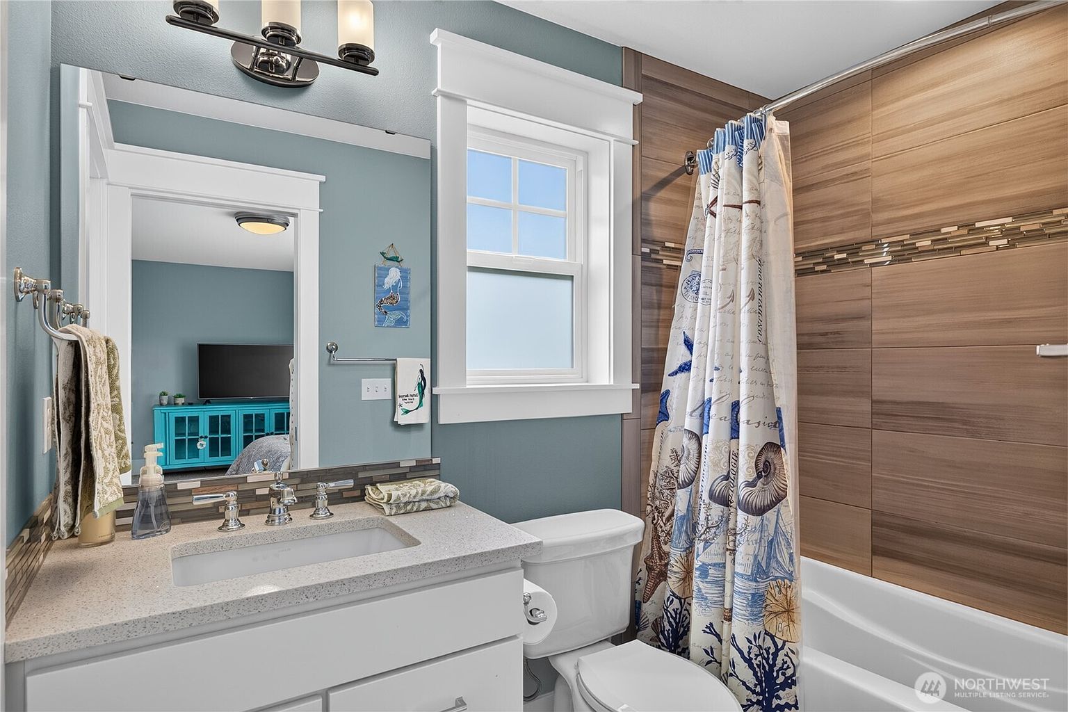 This bright bathroom features a white vanity with a light-colored stone countertop and a rectangular undermount sink. The shower area is accented with horizontal wood-look tiles and a decorative mosaic border, complemented by a nautical-themed shower curtain. A large mirror reflects the adjacent room, while a window above the toilet provides natural light to the blue-walled space.