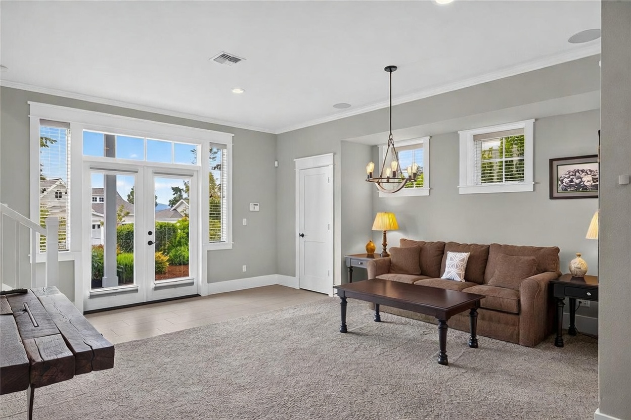 This bright and inviting living room features neutral gray walls, plush carpeting, and a comfortable brown sofa arranged around a dark wood coffee table. Large glass-paned double doors provide ample natural light and a view of the outdoor greenery, while a classic chandelier adds a touch of elegance to the space. The room is accented by a table lamp and a framed piece of wall art, creating a cozy and well-lit atmosphere.
