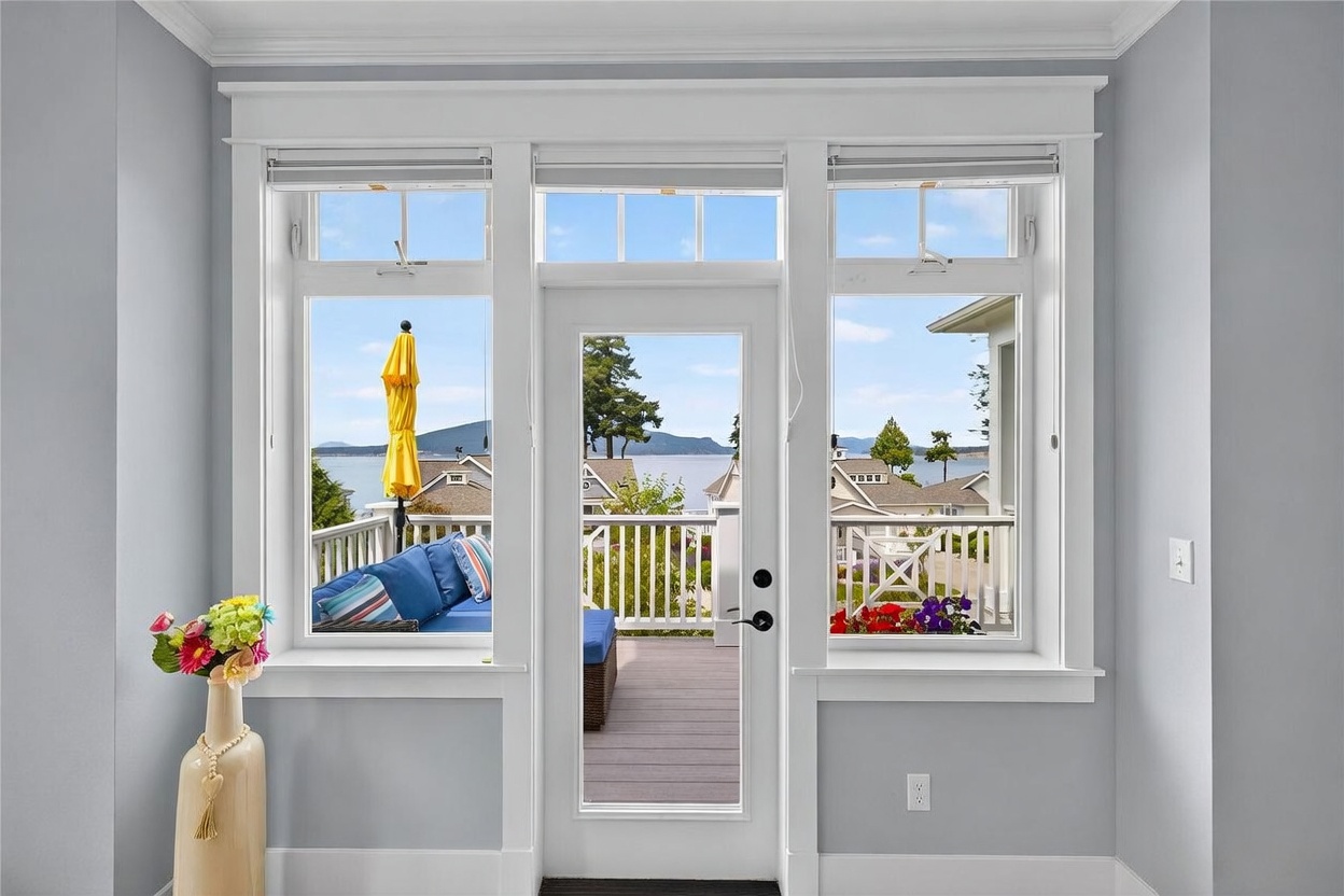 This interior view features a central glass-paned door flanked by two large windows, creating a bright and airy focal point that frames a scenic water view. The room is finished with light gray walls and crisp white trim, complemented by a decorative vase with colorful flowers on the left. Through the glass, a wooden deck with outdoor furniture is visible, seamlessly connecting the indoor living space to the exterior environment.