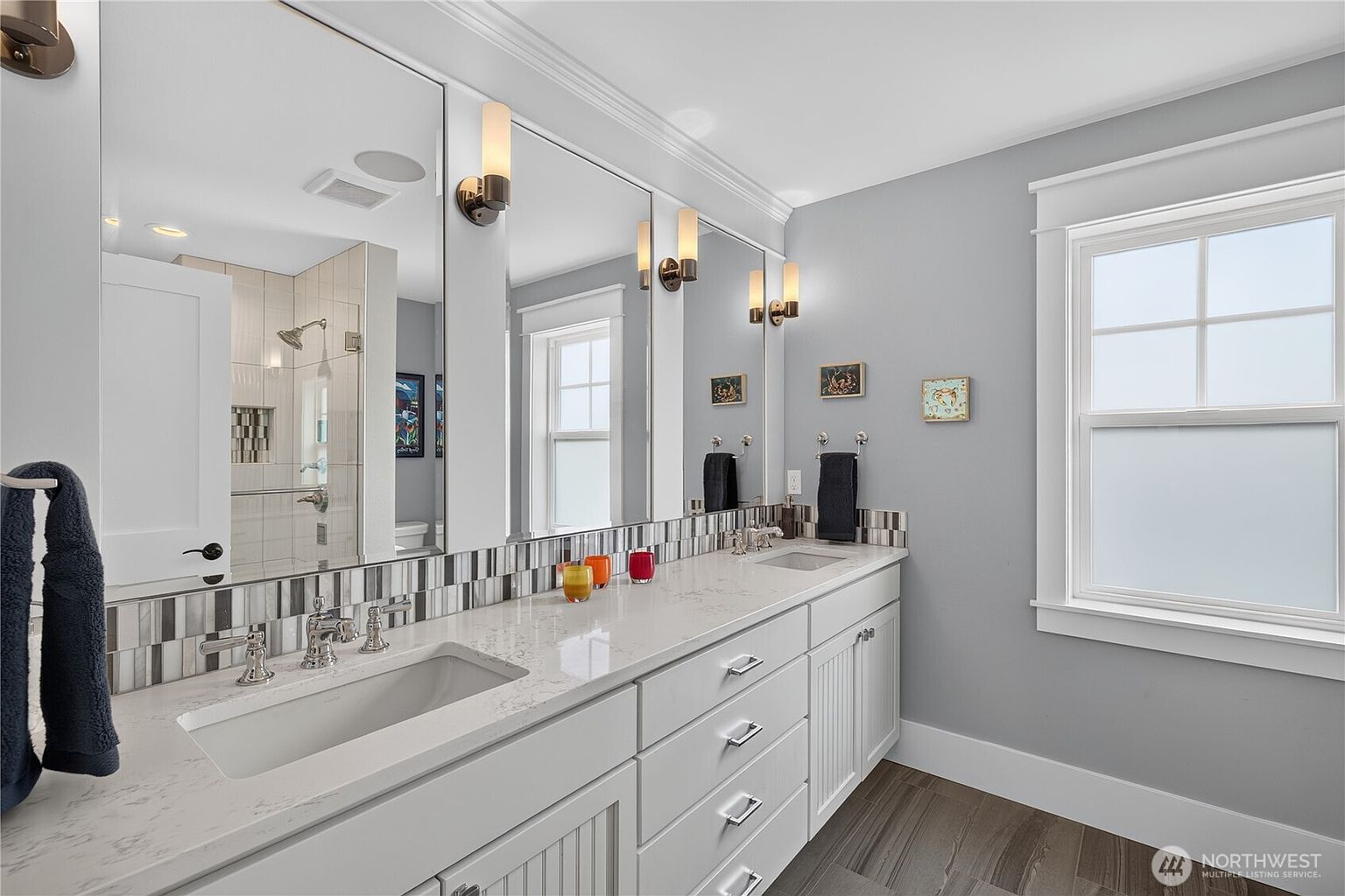 This bright and clean bathroom features a long dual-sink vanity with white cabinetry and a light-colored stone countertop. A decorative mosaic tile backsplash runs along the wall, complemented by large mirrors and wall-mounted light fixtures. The space includes a window with frosted glass for privacy and a view into the adjacent tiled shower area.