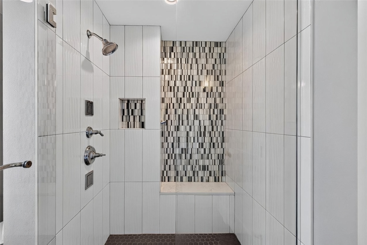 This modern walk-in shower features large-format white wall tiles contrasted by a striking vertical accent strip of mosaic tiles. The space includes a built-in bench with a stone top, a rainfall showerhead, and a handheld sprayer, all finished in polished chrome. A glass door provides a clear view into the shower, which is anchored by dark, hexagonal floor tiles.