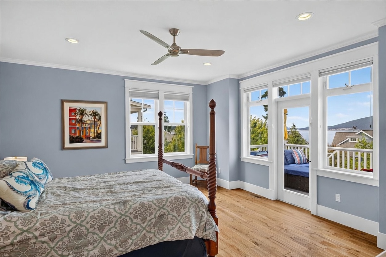 This bright and airy primary bedroom features light blue walls, warm-toned wood flooring, and a classic four-poster bed with a patterned duvet. Large windows and a glass door provide ample natural light and offer a scenic view of the water and surrounding landscape. A ceiling fan and recessed lighting complete the space, which opens directly onto a private balcony.