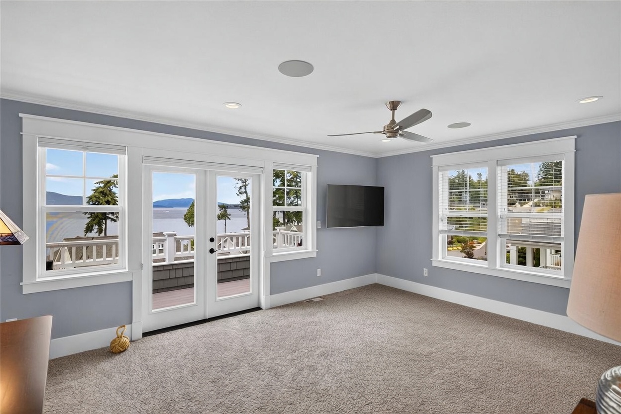 This primary bedroom features a serene, light blue color palette and plush carpeting, creating a calm and inviting atmosphere. A large set of French doors opens onto a deck with expansive water views, while additional windows provide ample natural light. The room is equipped with a ceiling fan and a wall-mounted television, offering a comfortable and functional living space.