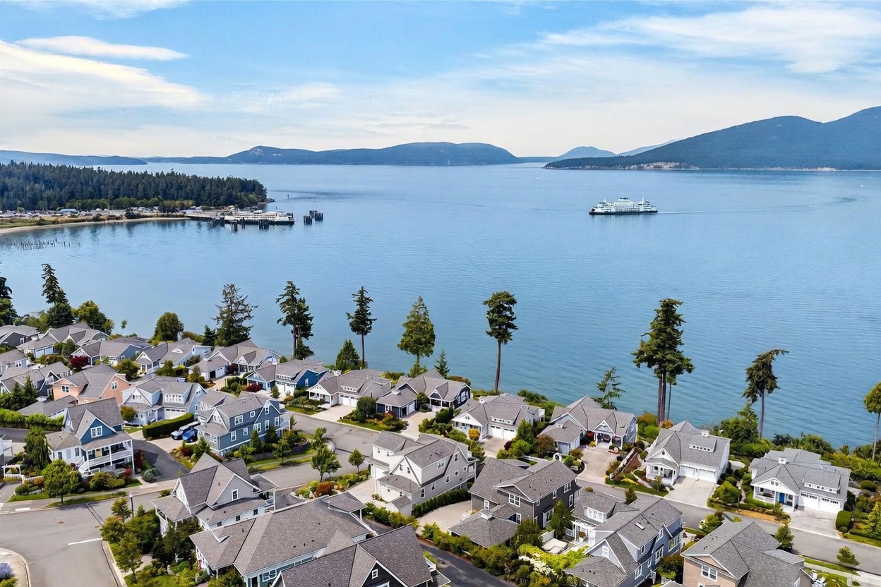 This high-angle aerial view captures a coastal residential neighborhood nestled along a scenic shoreline. The scene features a cluster of suburban homes with varied rooflines and neutral siding, set against the backdrop of a vast, calm bay with a ferry crossing in the distance. The composition emphasizes the community's proximity to the water, lush green trees, and the expansive horizon.
