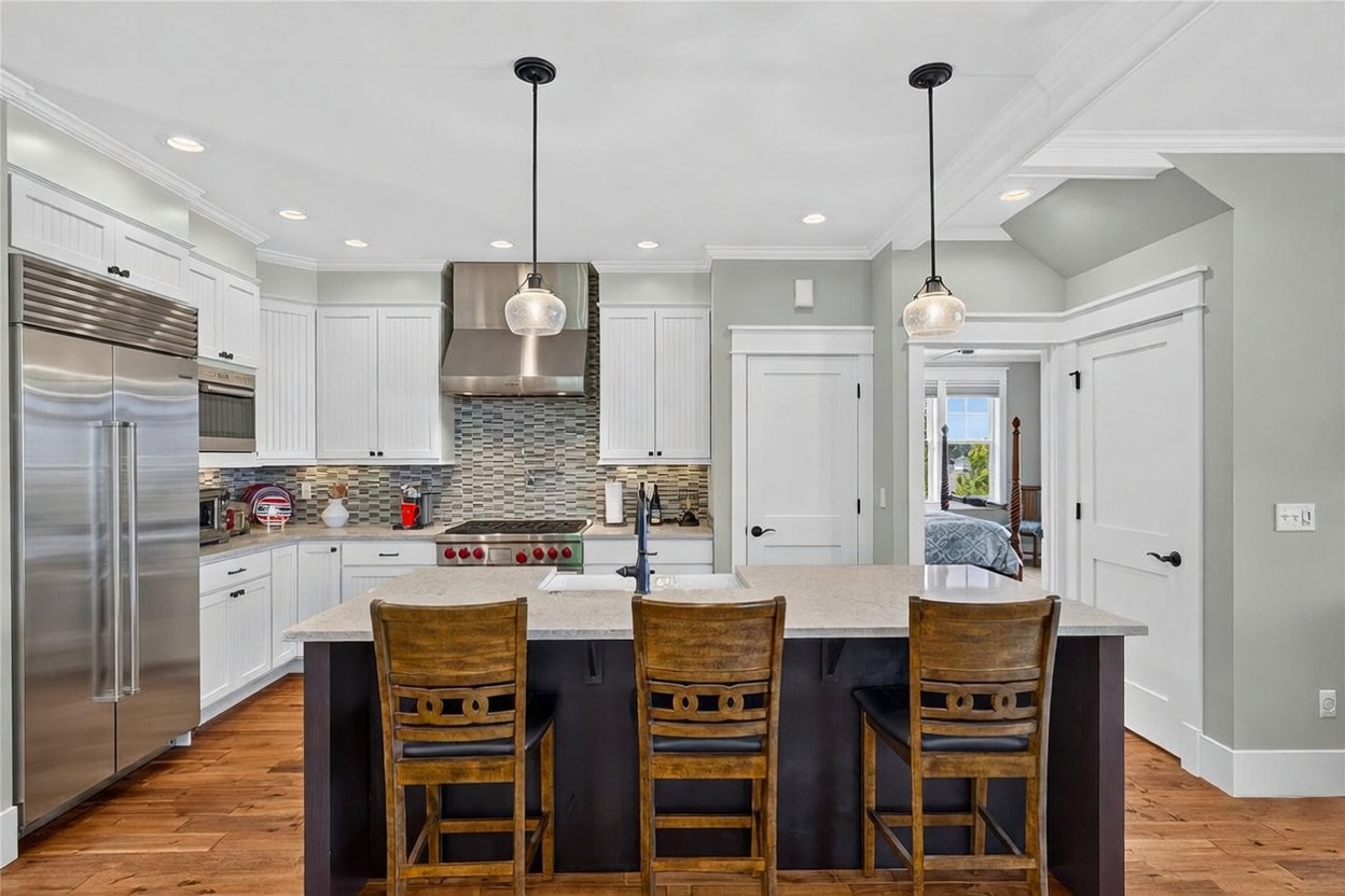 This bright and modern kitchen features white cabinetry, a stainless steel refrigerator, and a central island with dark base cabinetry and three wooden bar stools. A mosaic tile backsplash sits behind the stainless steel range and vent hood, while warm-toned wood flooring runs throughout the space. The perspective is a straight-on shot from the kitchen island, looking toward the cooking area and a doorway that reveals a glimpse of a bedroom.