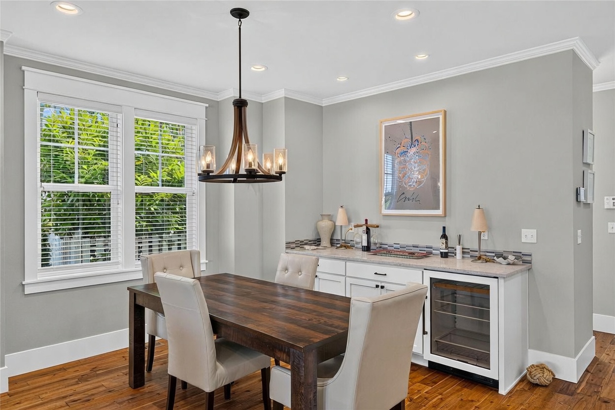 This dining area features a dark wood table paired with four upholstered chairs, illuminated by a rustic-style chandelier. To the side, a built-in wet bar includes white cabinetry, a stone countertop, a tiled backsplash, and a dedicated wine refrigerator. The room is finished with light gray walls, crown molding, and large windows that allow for ample natural light.