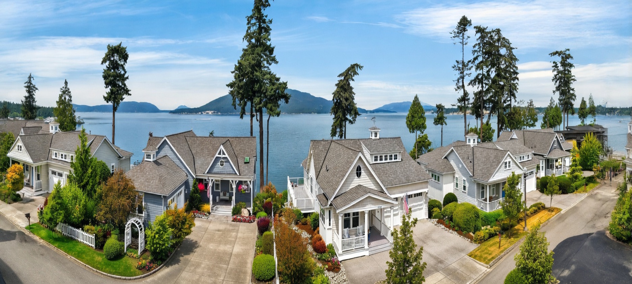 This panoramic view captures a row of charming, coastal-style homes situated along a waterfront, showcasing their front facades and manicured landscaping. The houses feature classic architectural details like gabled roofs, covered porches, and shingle-style siding, all set against a backdrop of a serene bay and distant mountains. The perspective provides a wide, cinematic look at the neighborhood's inviting curb appeal and its prime location near the water.