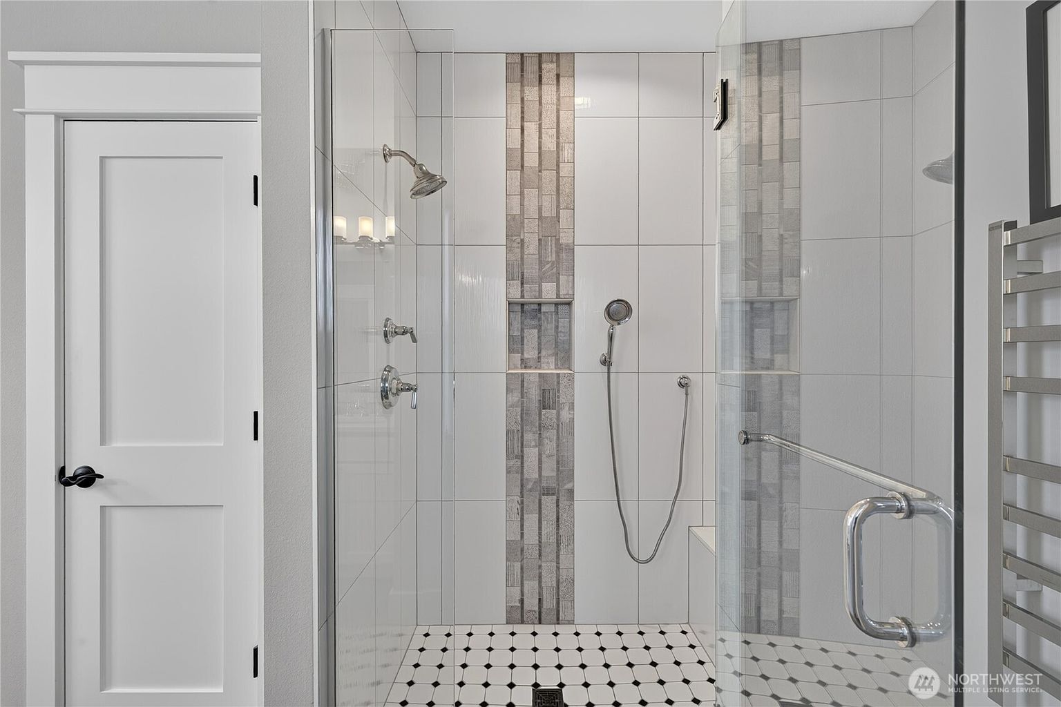 This modern walk-in shower features a glass enclosure and a striking vertical accent strip of mosaic tile centered on the back wall. The shower includes both a fixed showerhead and a handheld sprayer, along with a built-in recessed niche for storage and a small bench. The floor is finished with a classic black-and-white patterned tile, creating a clean and sophisticated aesthetic.