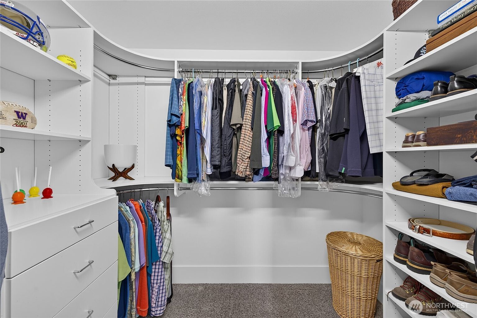 This organized walk-in closet features white custom shelving and hanging rods arranged in a curved layout to maximize storage space. The room includes a built-in dresser with drawers, open shelving for accessories, and a dedicated area for hanging garments, all set against a neutral backdrop with carpeted flooring. A woven basket sits on the floor, adding a touch of texture to the clean, functional design.