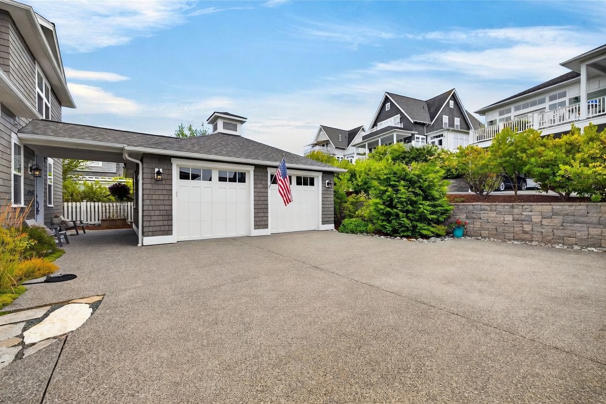 This image captures a detached two-car garage with gray shingle siding and a covered breezeway connecting to the main house. A paved driveway leads up to the white garage doors, which are adorned with an American flag, while a stone retaining wall and lush landscaping frame the elevated backyard area. The perspective is a wide-angle shot from the driveway, showcasing the property's curb appeal and well-maintained exterior.