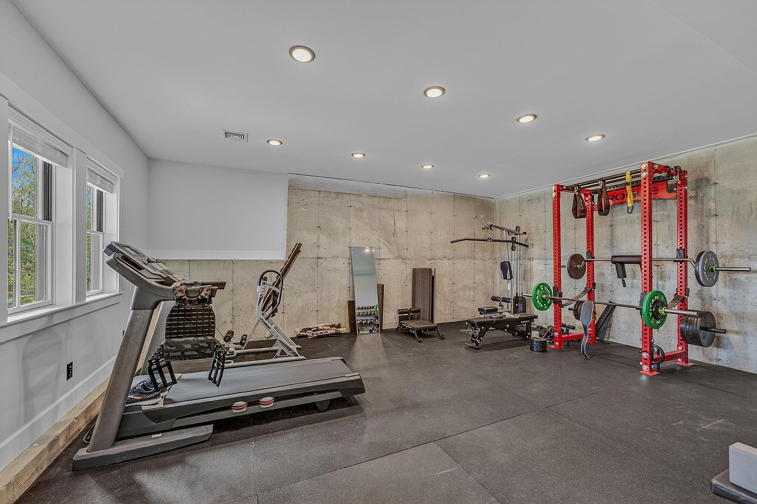 This spacious home gym features durable rubber flooring and exposed concrete walls, creating an industrial and functional workout environment. The room is equipped with a professional-grade power rack, a treadmill, and various strength training accessories, all illuminated by recessed ceiling lighting. Large windows on the left side provide natural light, making the space feel open and well-suited for a dedicated fitness routine.