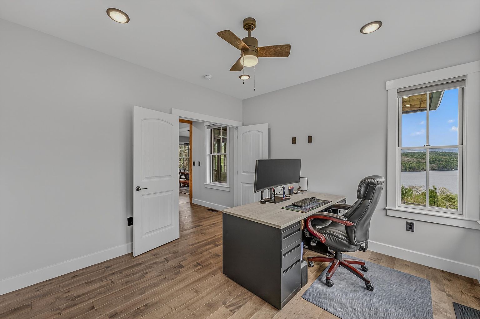 This bright home office features light gray walls, wood-look flooring, and a modern desk positioned to face the room. A large window provides a scenic view of a lake and surrounding trees, while a ceiling fan with integrated lighting hangs above. The room includes a doorway leading to an adjacent space, creating an open and functional workspace environment.