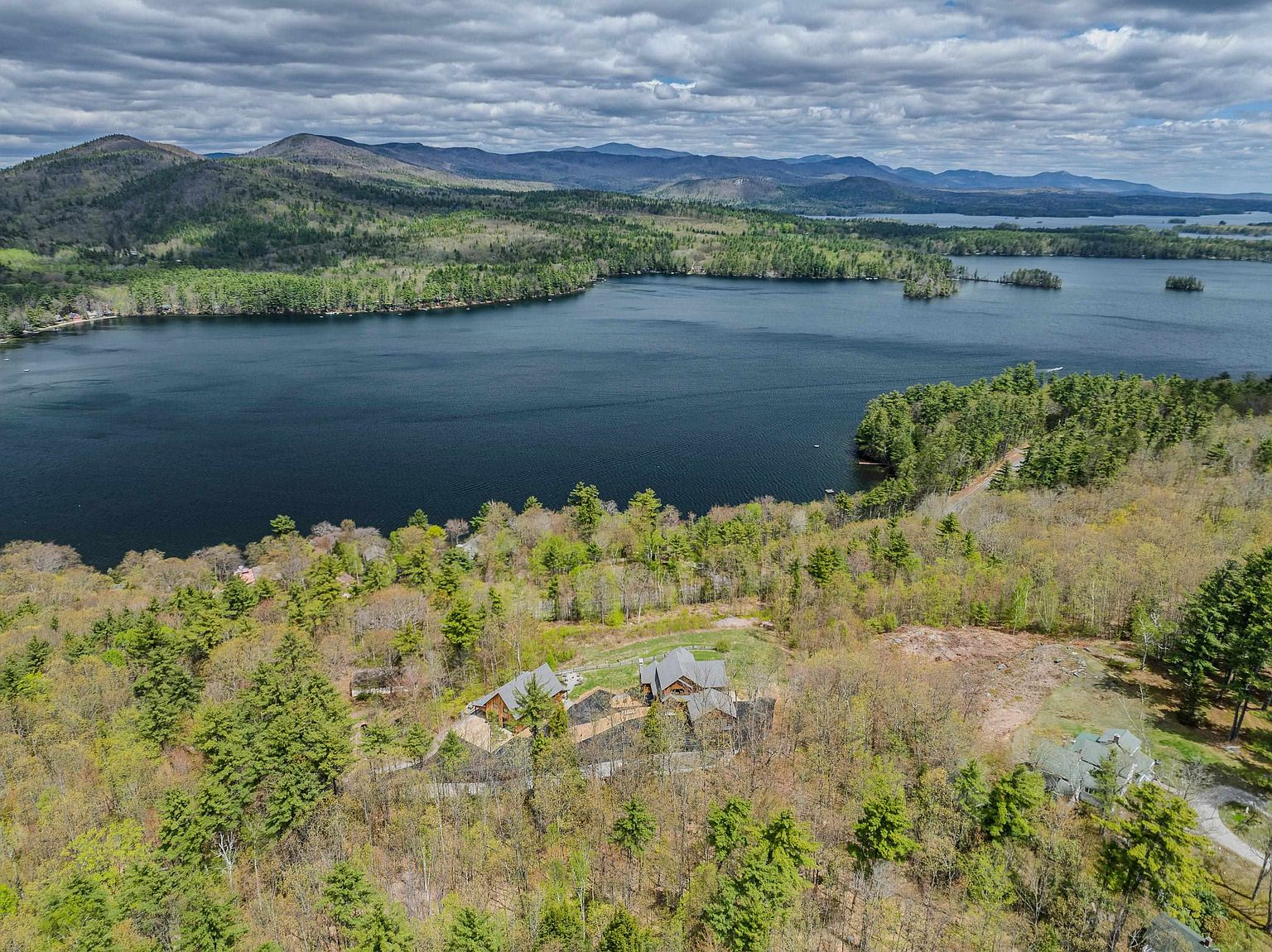 This high-angle aerial perspective captures a sprawling property nestled within a dense, lush forest overlooking a large, serene lake. The main residence features a complex, multi-gabled roofline and is surrounded by mature trees, with a secondary structure visible nearby. The expansive landscape stretches toward distant mountain ranges under a dramatic, cloud-filled sky, emphasizing the property's secluded and scenic location.