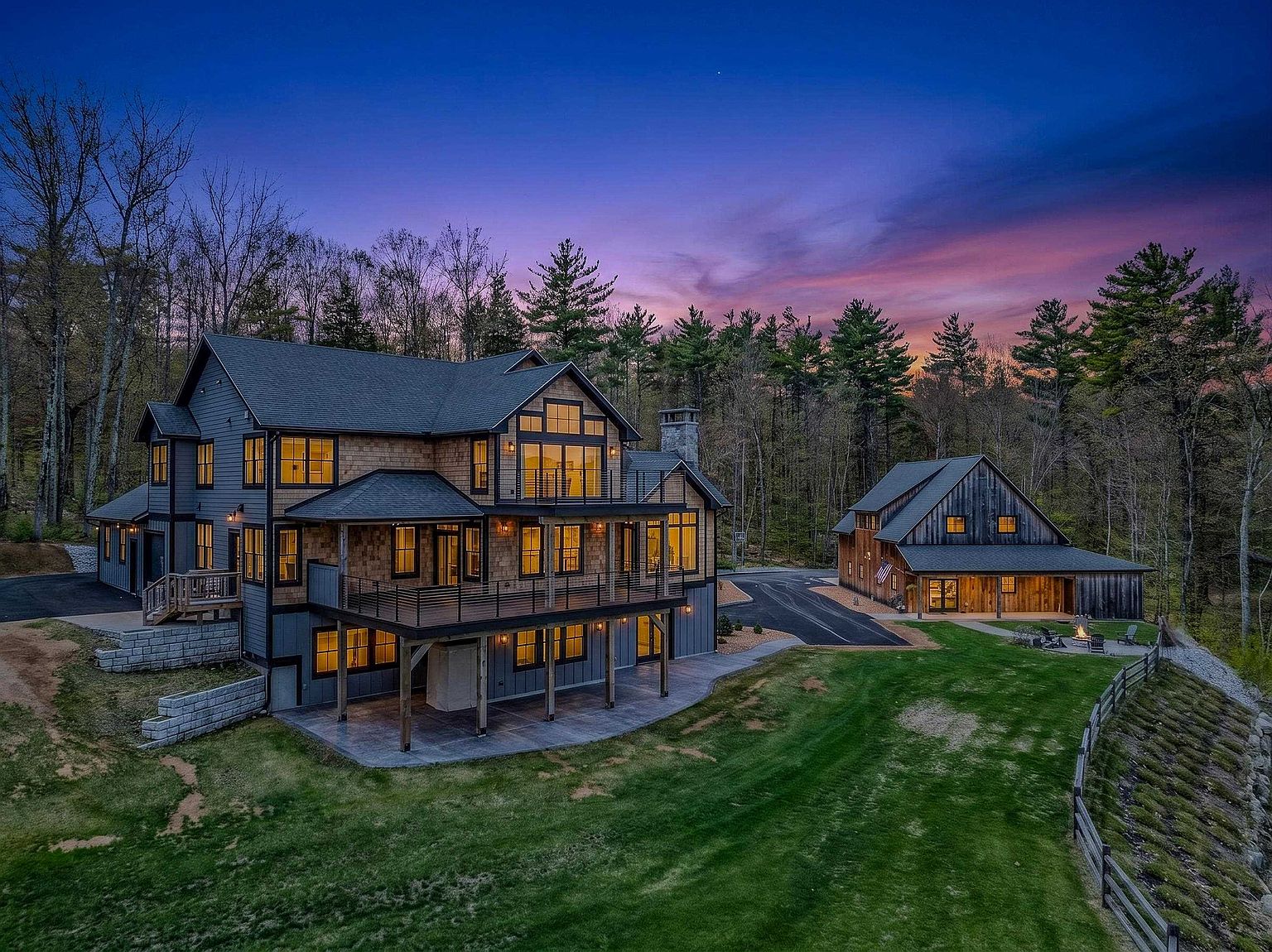 This aerial perspective captures a sprawling, multi-level custom home and a detached barn-style structure set against a vibrant sunset sky. The main residence features a blend of dark siding and natural wood shingles, with expansive decks and balconies overlooking a manicured lawn and surrounding forest. The scene highlights the property's secluded, wooded setting and the cohesive architectural design of the two primary buildings.