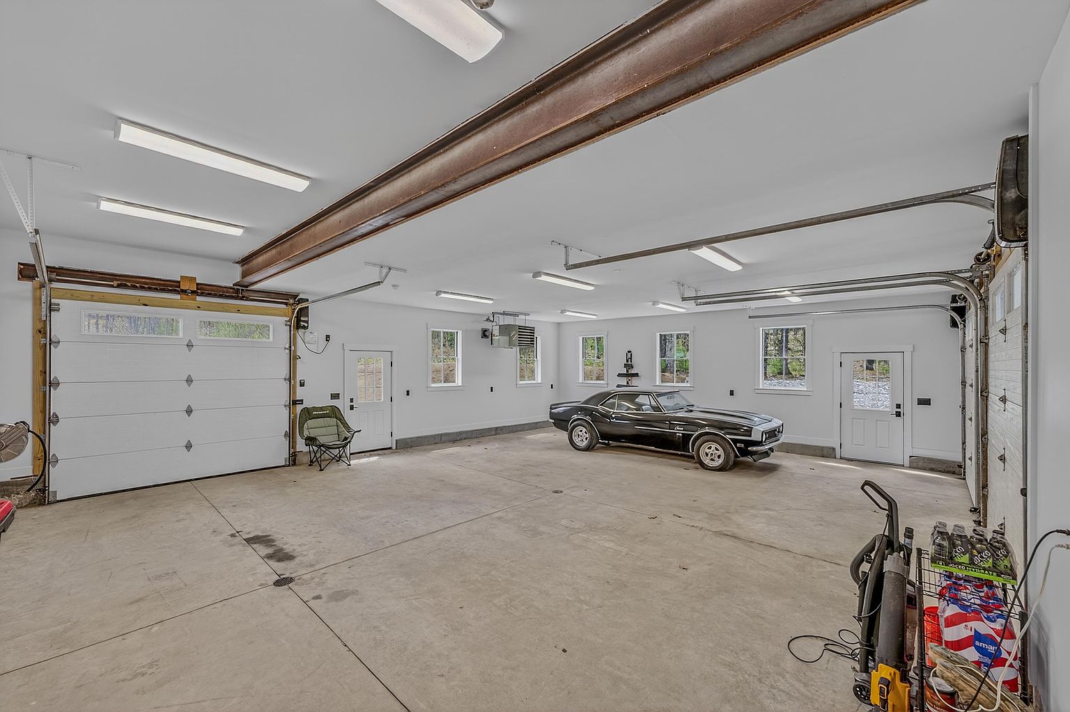 This spacious, well-lit garage features a clean concrete floor and white walls, providing a bright and organized environment. A classic black muscle car is parked in the center, while a folding chair sits near the side door, emphasizing the versatile nature of the space. The room is equipped with multiple overhead light fixtures and a prominent steel support beam running across the ceiling, highlighting the sturdy construction of this functional area.