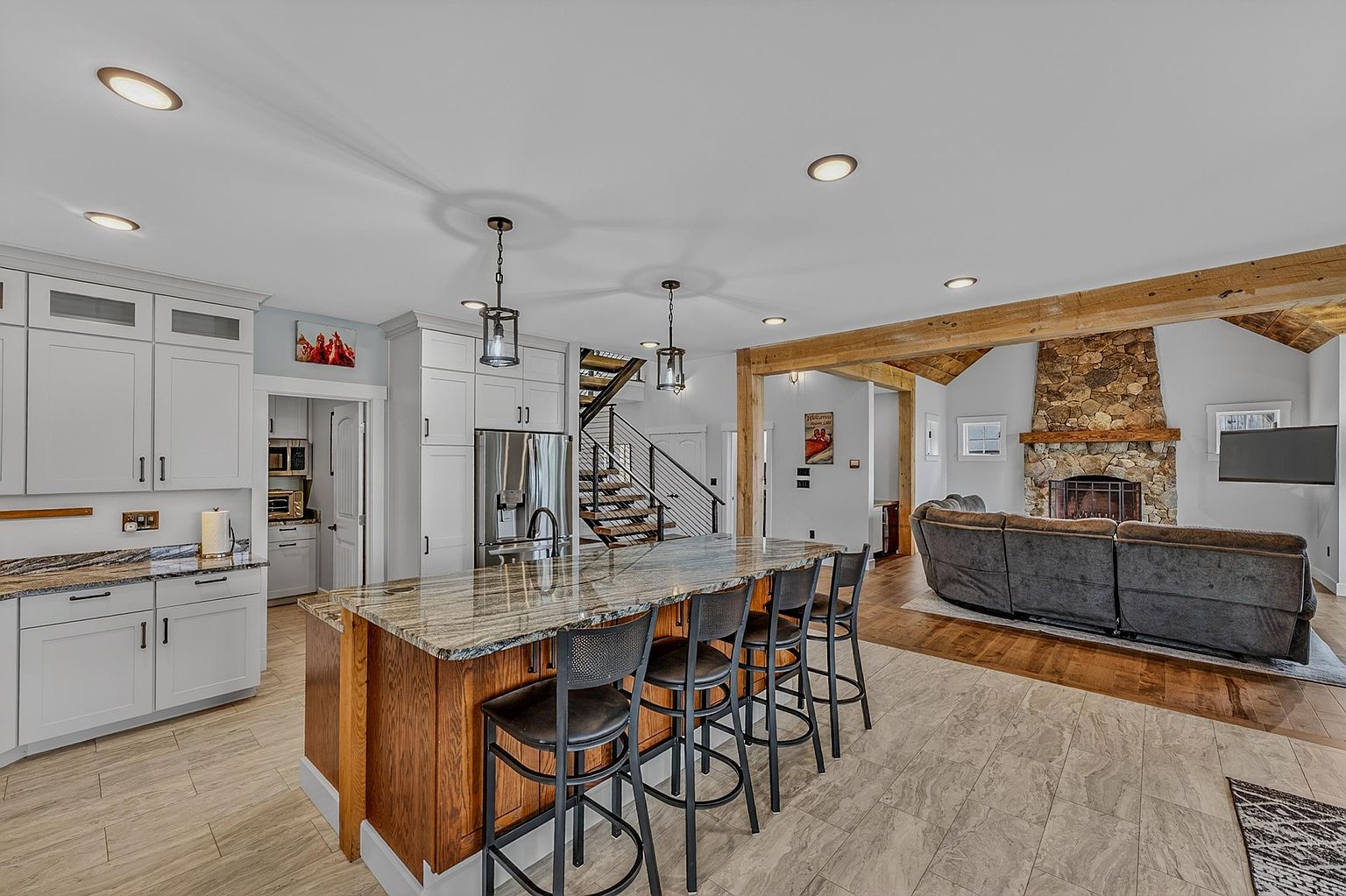 This open-concept kitchen features white cabinetry paired with a large central island topped with patterned stone and supported by a warm-toned wood base. The space flows seamlessly into a living area defined by a prominent stone-clad chimney breast and exposed wooden beams. The perspective captures the transition between the tiled kitchen floor and the hardwood living room, highlighting the home's spacious and inviting layout.
