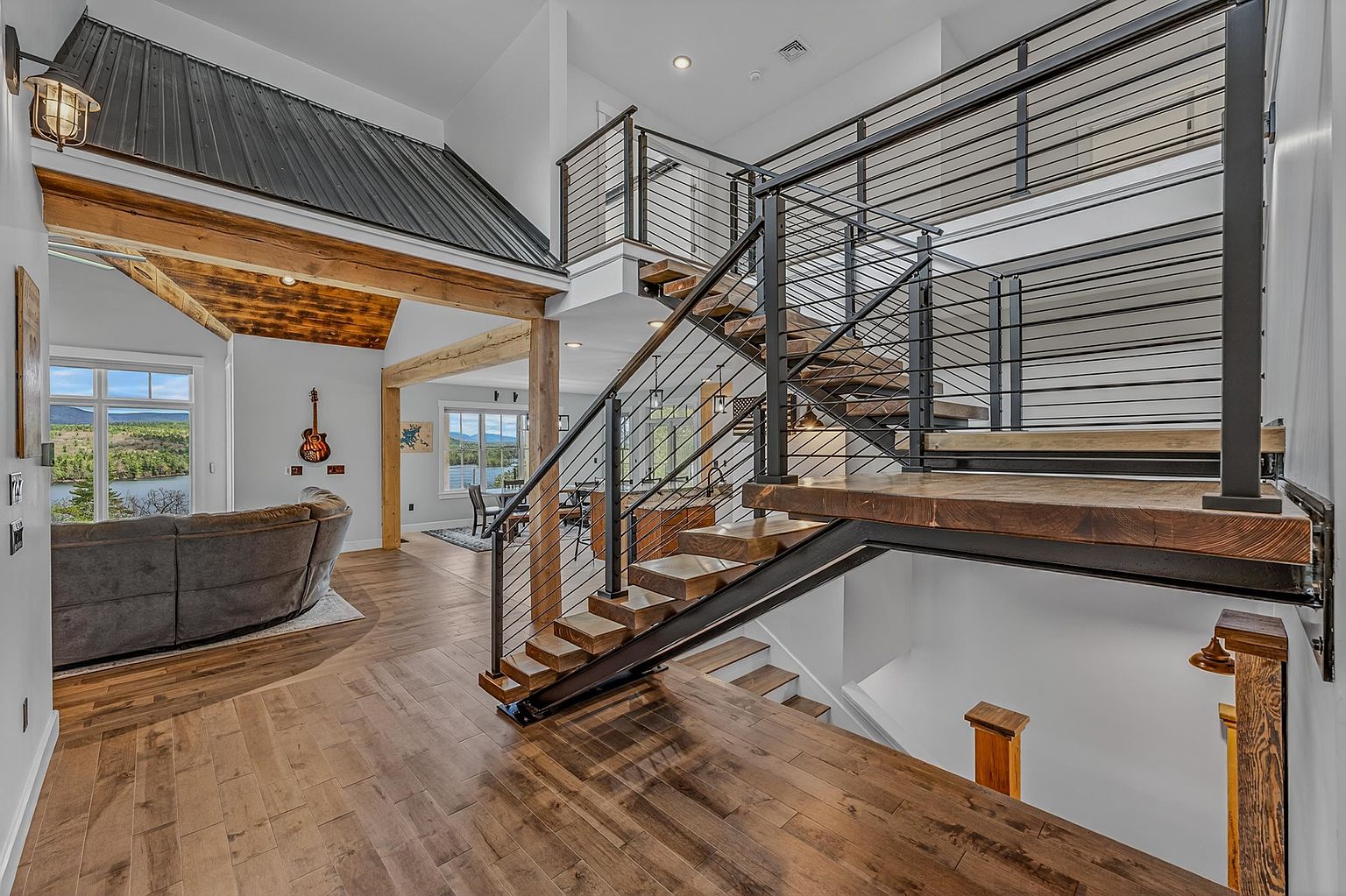 This interior view highlights a striking open-concept staircase featuring dark metal railings with horizontal cable infill and thick, warm-toned wood treads. The space showcases a blend of rustic and industrial design, with exposed timber beams, a metal-clad vaulted ceiling section, and rich wood flooring throughout. The perspective looks from the landing toward an open living area and kitchen, emphasizing the airy, multi-level layout and the scenic outdoor views visible through large windows.