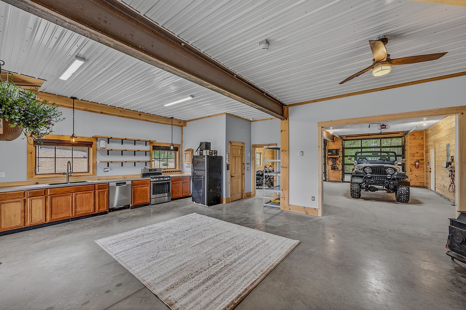 This spacious, industrial-style interior features a functional kitchen area integrated into a large, open-concept garage space with concrete floors. The kitchen is equipped with wooden cabinetry, stainless steel appliances, and open shelving, while the adjacent bay provides a clear view of a parked Jeep. The room is illuminated by overhead lighting and a ceiling fan, creating a versatile, multi-purpose environment that blends utility with living space.