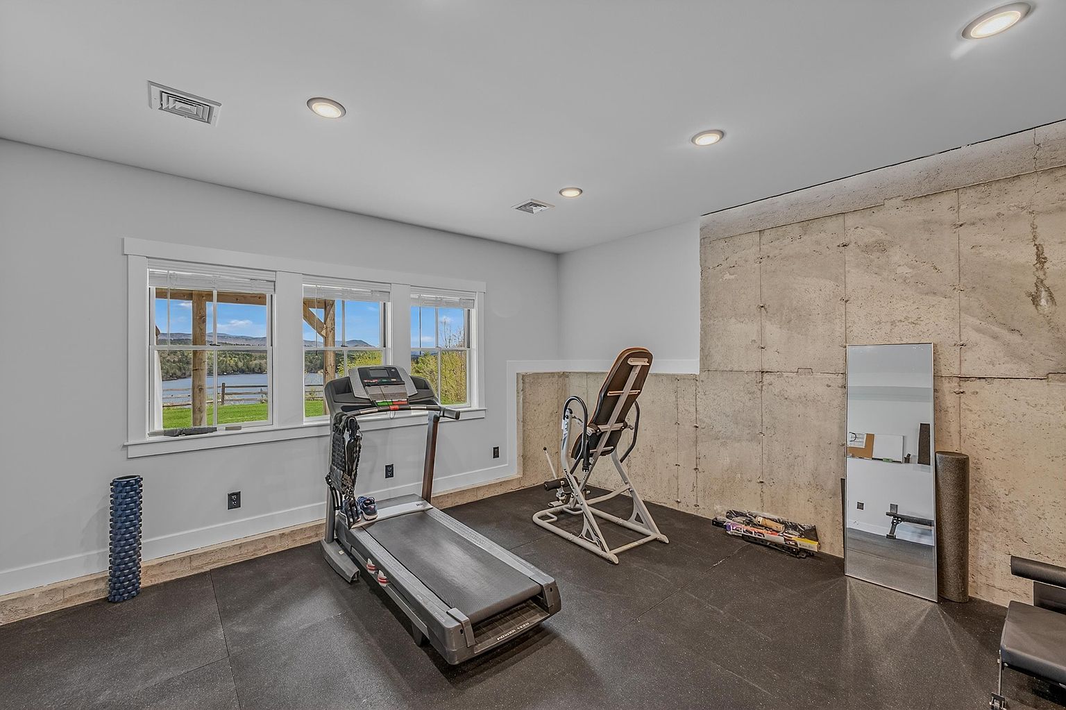 This home gym features a durable black rubber floor and a prominent raw concrete wall that adds an industrial aesthetic to the space. A treadmill and an inversion table are positioned in the center, while a tall floor mirror leans against the concrete wall. Large windows provide ample natural light and a scenic view of the lake and surrounding landscape.