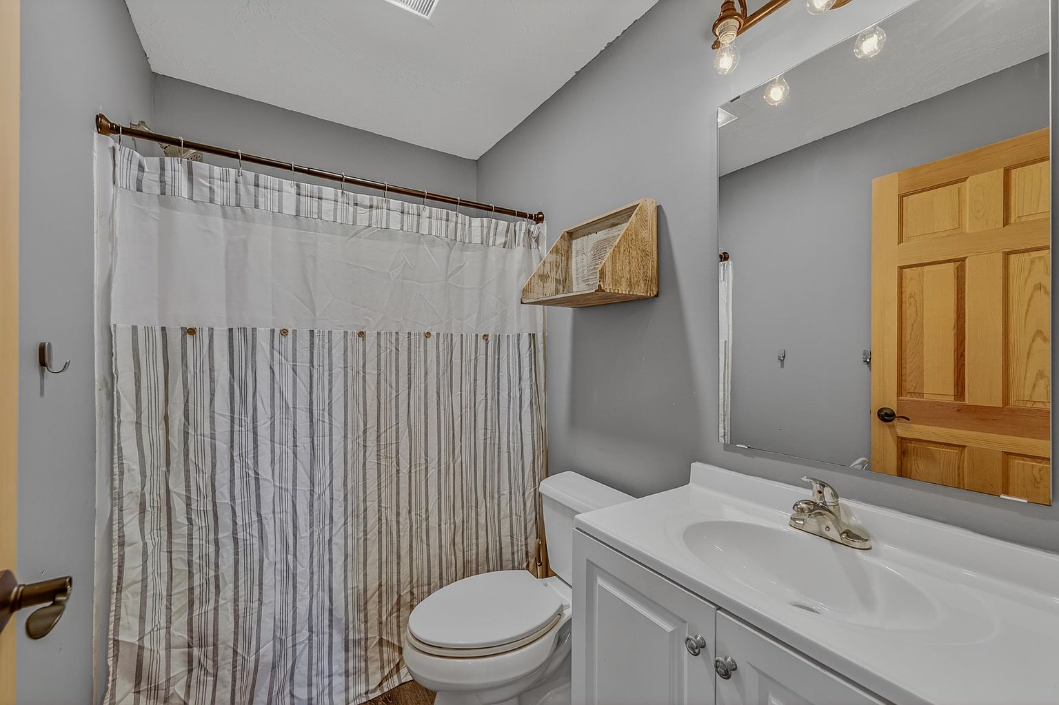 This compact bathroom features a white vanity with a single sink, a toilet, and a shower area enclosed by a striped curtain. A large mirror above the vanity reflects the wooden door, while a rustic wooden shelf is mounted on the gray-painted wall. The space is illuminated by a multi-bulb light fixture, creating a functional and neutral aesthetic.