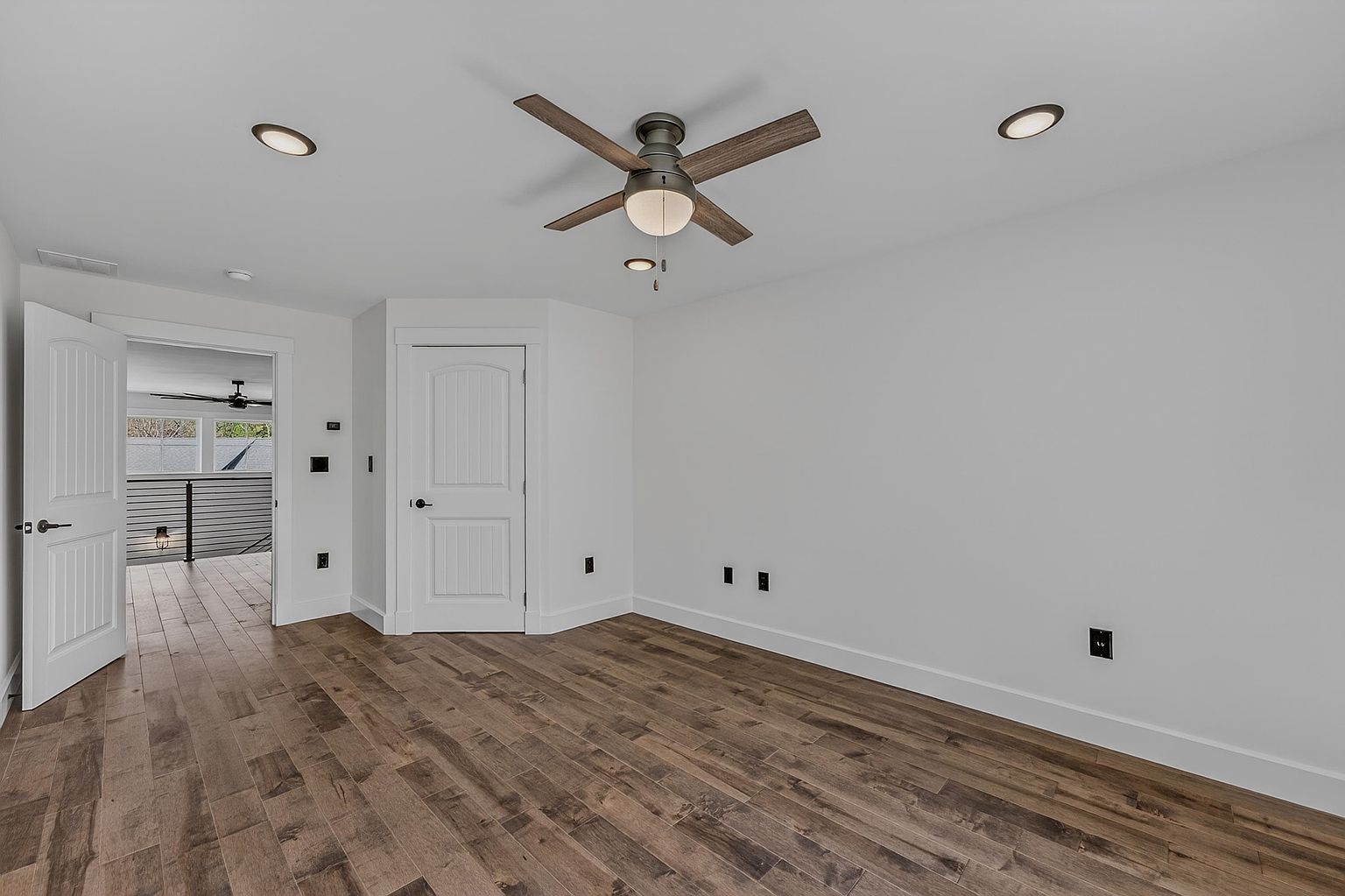This bright, neutral bedroom features wide-plank wood-look flooring and crisp white walls, creating a clean and versatile living space. A central ceiling fan with integrated lighting provides both air circulation and illumination, complemented by recessed lighting. The room includes a single interior door and an open doorway leading to a hallway with a metal-railed staircase, offering a sense of depth and architectural flow.