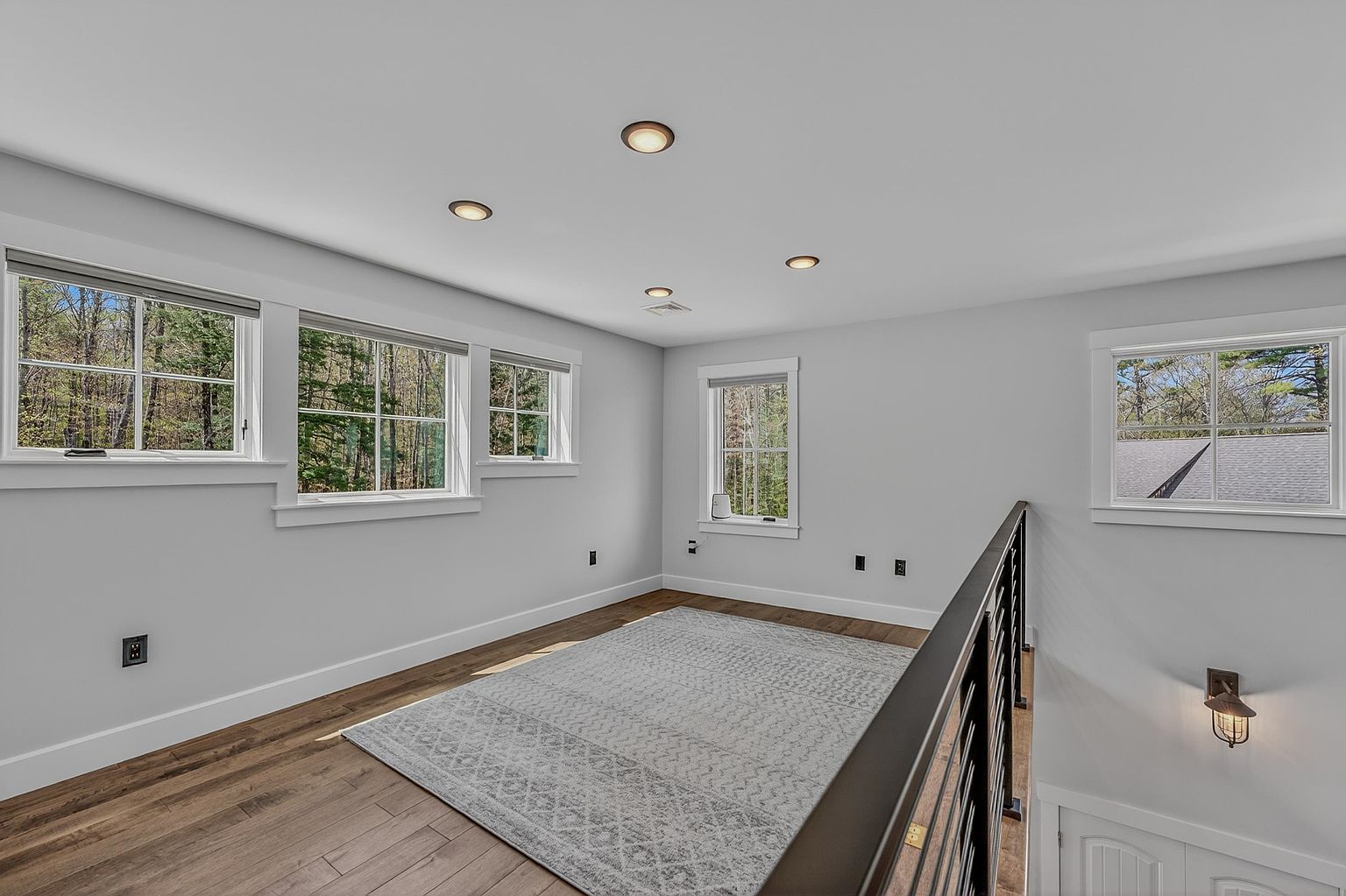 This bright lofted landing area features light-toned wood flooring and a neutral color palette, creating an airy and open atmosphere. A large, patterned area rug anchors the space, which is illuminated by recessed ceiling lights and natural light streaming through multiple windows. The area is bordered by a dark metal railing that overlooks the lower level, providing a modern architectural touch to the home's interior.