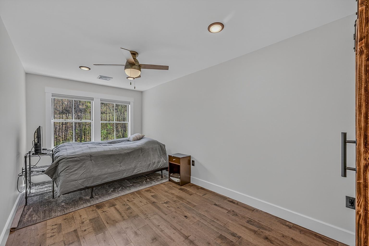 This bedroom features light-toned walls and warm-toned wood-look flooring, creating a bright and neutral atmosphere. A single bed with a grey comforter is positioned against a window wall, accompanied by a small wooden nightstand and a metal media stand. The room is illuminated by a ceiling fan with integrated lighting and recessed lights, offering a clean and functional space.