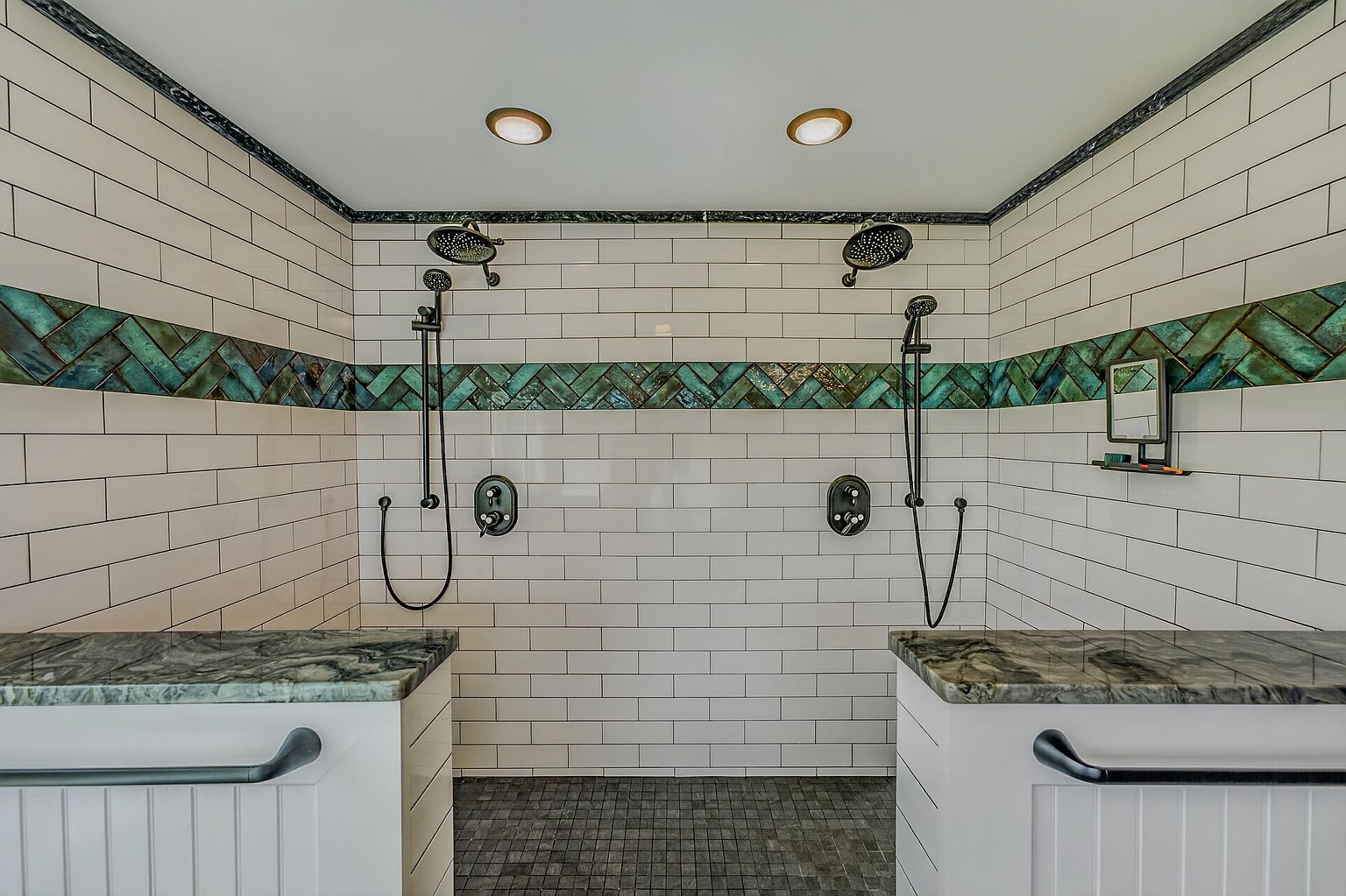 This spacious walk-in shower features dual shower heads and a central tiled wall with a decorative green herringbone-patterned accent band. The space is framed by two white-paneled vanity bases topped with dark, veined stone counters and equipped with safety grab bars. The floor is finished in a dark, grid-patterned tile, creating a clean and functional aesthetic.