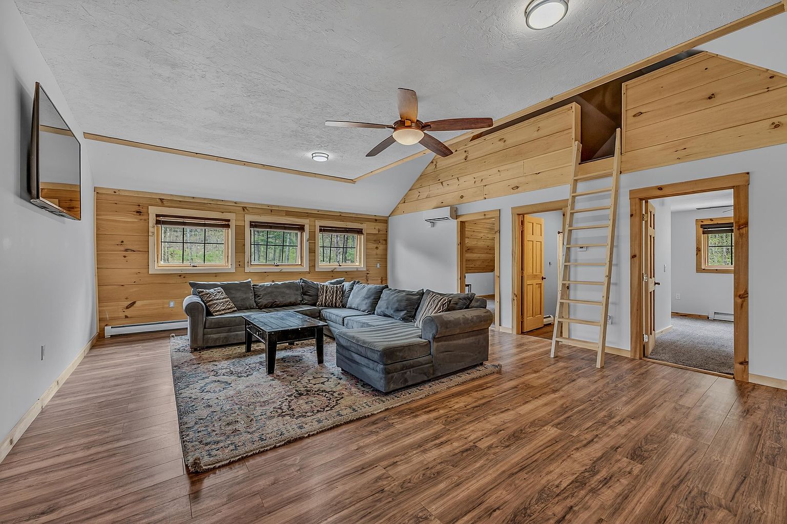 This spacious living room features a rustic aesthetic with light wood paneling on the walls and a vaulted ceiling. A large, grey sectional sofa sits atop a patterned area rug, facing a wall-mounted television, while a wooden ladder provides access to a lofted area above. The room is brightened by three windows and includes doorways leading to adjacent spaces, creating an open and inviting atmosphere.