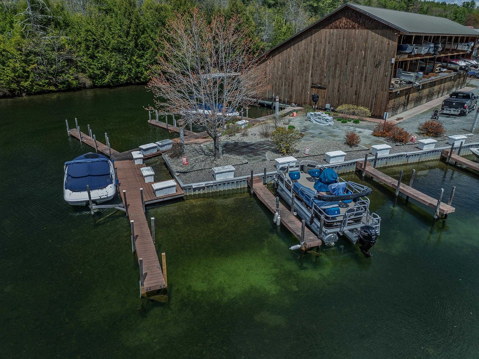 This aerial perspective captures a waterfront property featuring a large, rustic wooden boat storage building and a private dock system extending into clear, green-tinted water. Two boats are moored at the docks, which are connected by a stone-paved patio area shaded by a mature, budding tree. The scene conveys a peaceful, recreational lakeside atmosphere, highlighting the direct water access and functional outdoor storage facilities.
