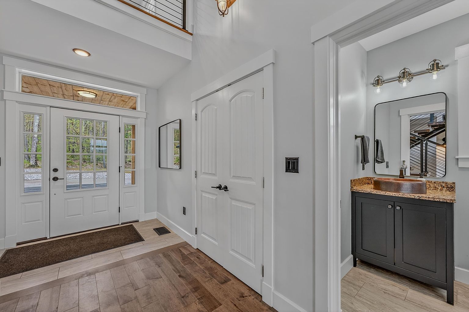 This bright entryway features a white front door with glass panes, leading into a foyer with warm-toned wood flooring. To the right, a doorway opens into a powder room equipped with a dark vanity, a copper-toned vessel sink, and a modern light fixture. The space is characterized by clean lines, neutral wall tones, and a welcoming, open-concept layout.