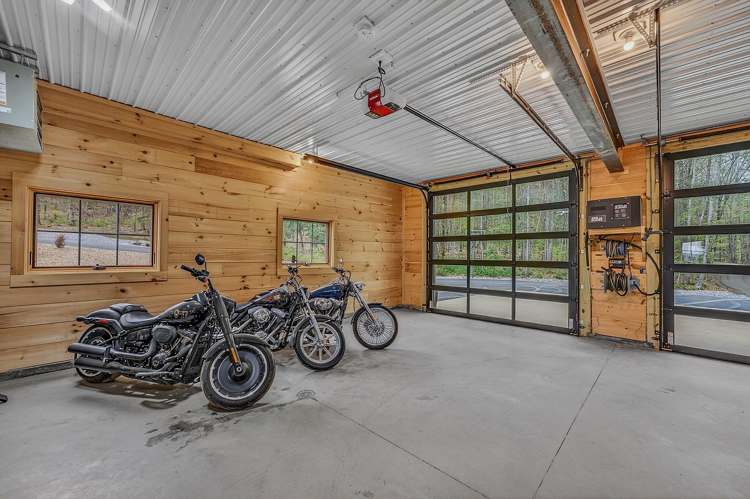 This spacious, well-lit garage features warm wood-paneled walls and a durable concrete floor, housing three motorcycles parked in a row. The space is equipped with a modern glass-paneled sectional garage door that provides a clear view of the wooded exterior, while a secondary matching door is visible to the right. The industrial-style ceiling with exposed metal beams and a mounted garage door opener adds to the functional, workshop-inspired aesthetic.