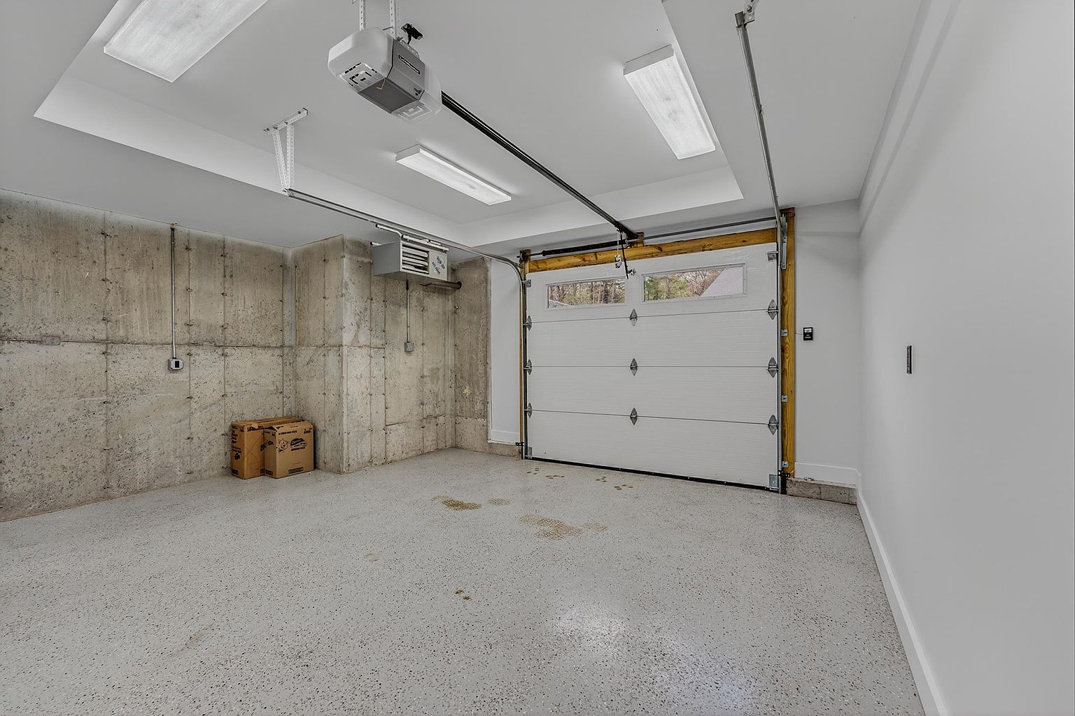 This image shows the interior of a clean, well-maintained garage featuring a white sectional door with windows and a speckled epoxy floor. The space includes exposed concrete walls, a ceiling-mounted garage door opener, and recessed lighting fixtures. Two cardboard boxes sit in the corner, and a wall-mounted heater is visible, suggesting a functional and climate-controlled environment.