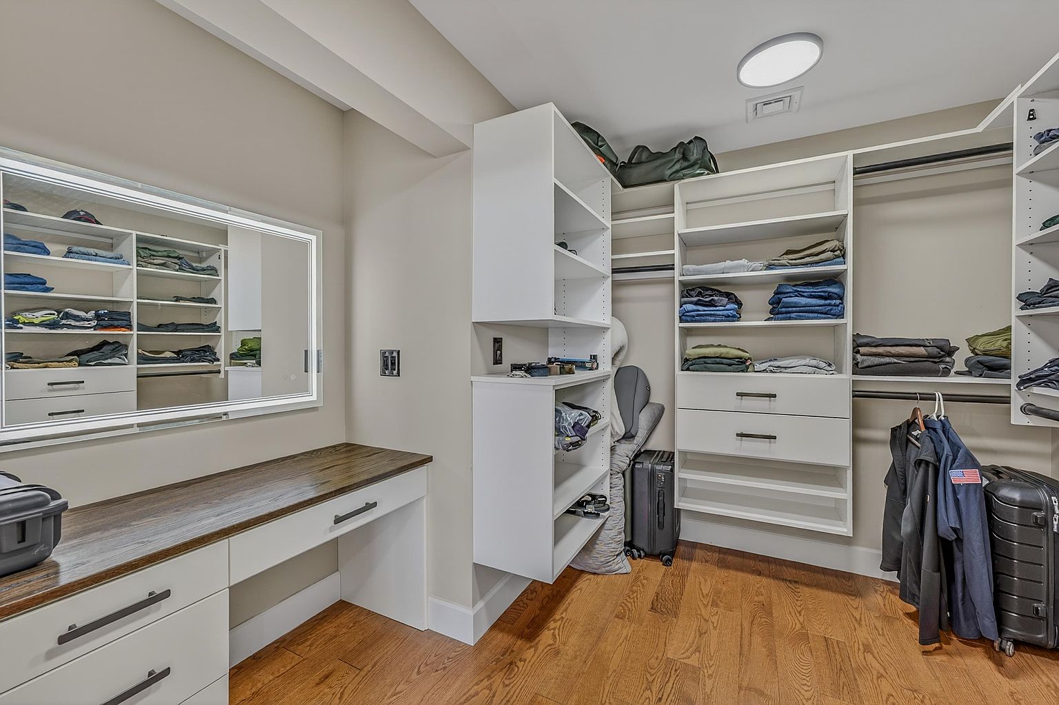 This organized walk-in closet features custom white shelving units, hanging rods, and a built-in vanity area with a dark wood-toned countertop. A large, illuminated mirror hangs above the vanity, while the room is finished with warm-toned wood flooring and recessed ceiling lighting. The space is efficiently designed to maximize storage with a mix of open shelving, drawers, and hanging space.