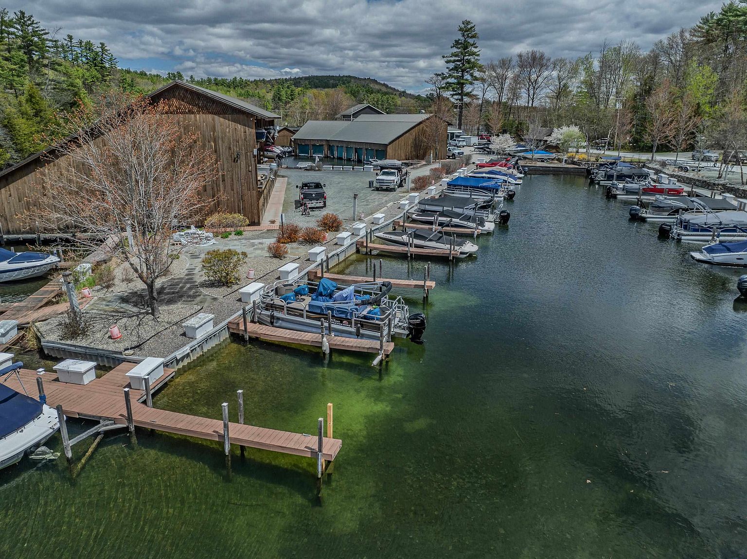 This aerial view captures a serene marina setting featuring a large wooden boat storage building and a series of docks extending into clear, green-tinted water. Numerous pontoon boats are moored along the docks, while a gravel parking area sits between the building and the water's edge. The scene is framed by lush trees and a cloudy sky, creating a peaceful and functional waterfront environment.