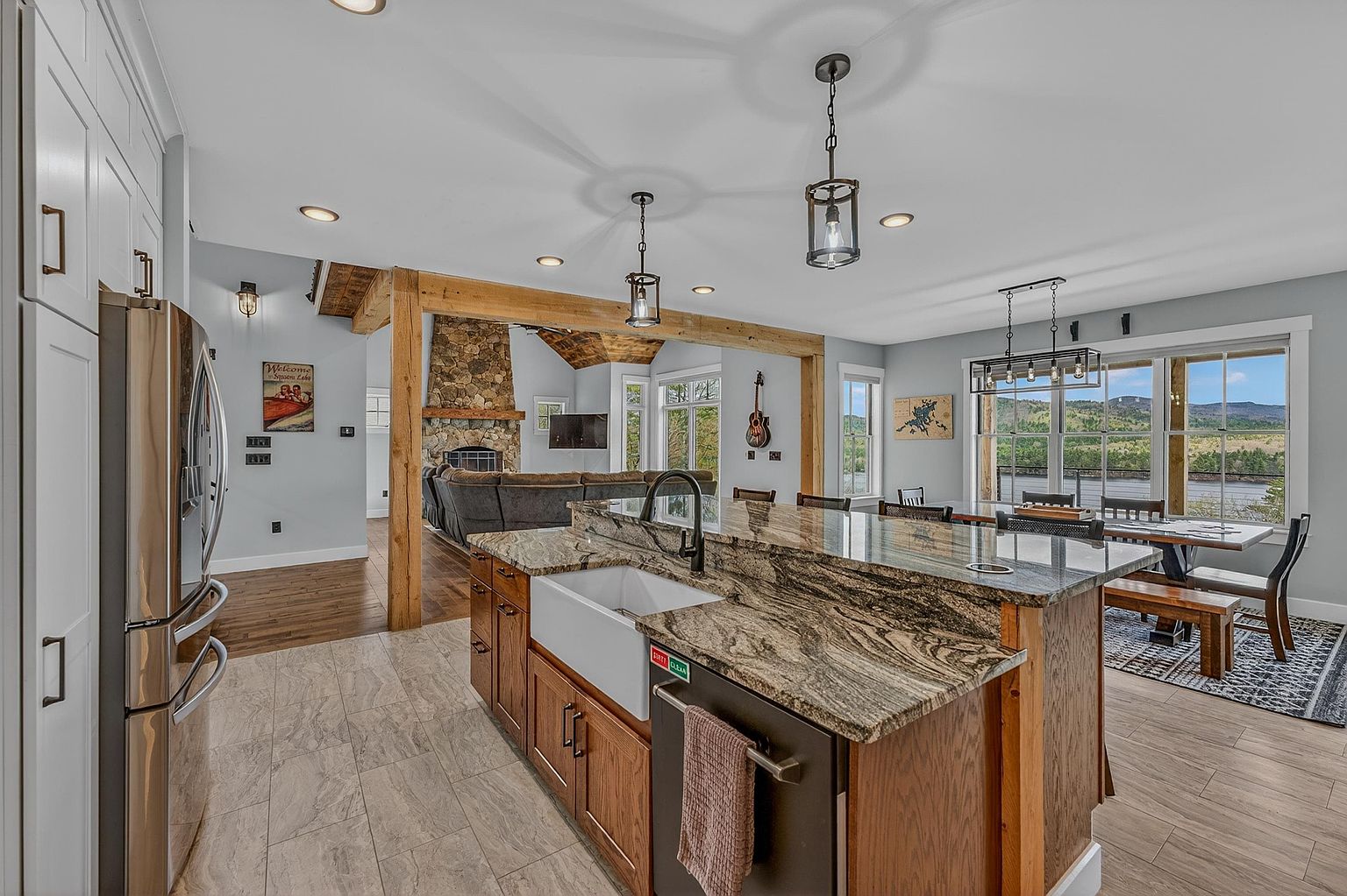 This open-concept kitchen features a large central island with a farmhouse sink and stone countertops, serving as the focal point of the space. The room flows seamlessly into a dining area with large windows overlooking a scenic landscape and a living area anchored by a prominent stone-clad chimney breast. Warm wood accents, pendant lighting, and a neutral color palette create a welcoming and cohesive atmosphere.