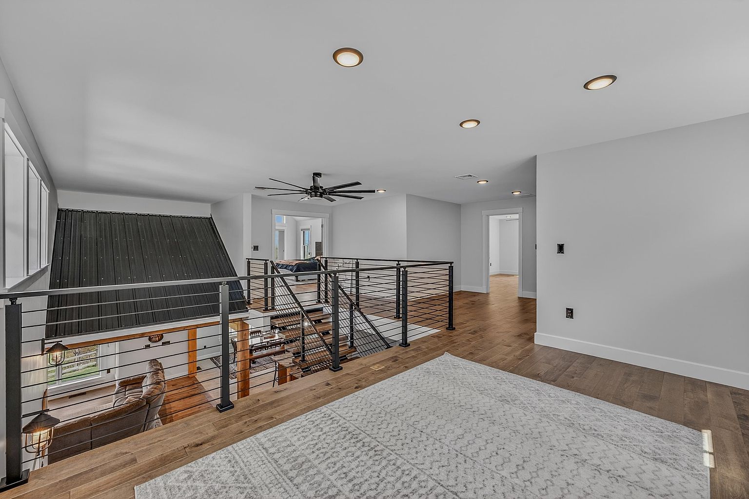 This image captures an open-concept loft area overlooking a lower living space, characterized by a modern industrial aesthetic. A sleek black metal railing with horizontal cables separates the upper landing from the staircase, while a large, light-colored area rug anchors the wooden floor. The perspective looks out from the landing toward a bedroom doorway, highlighting the airy, bright atmosphere created by recessed lighting and neutral wall tones.