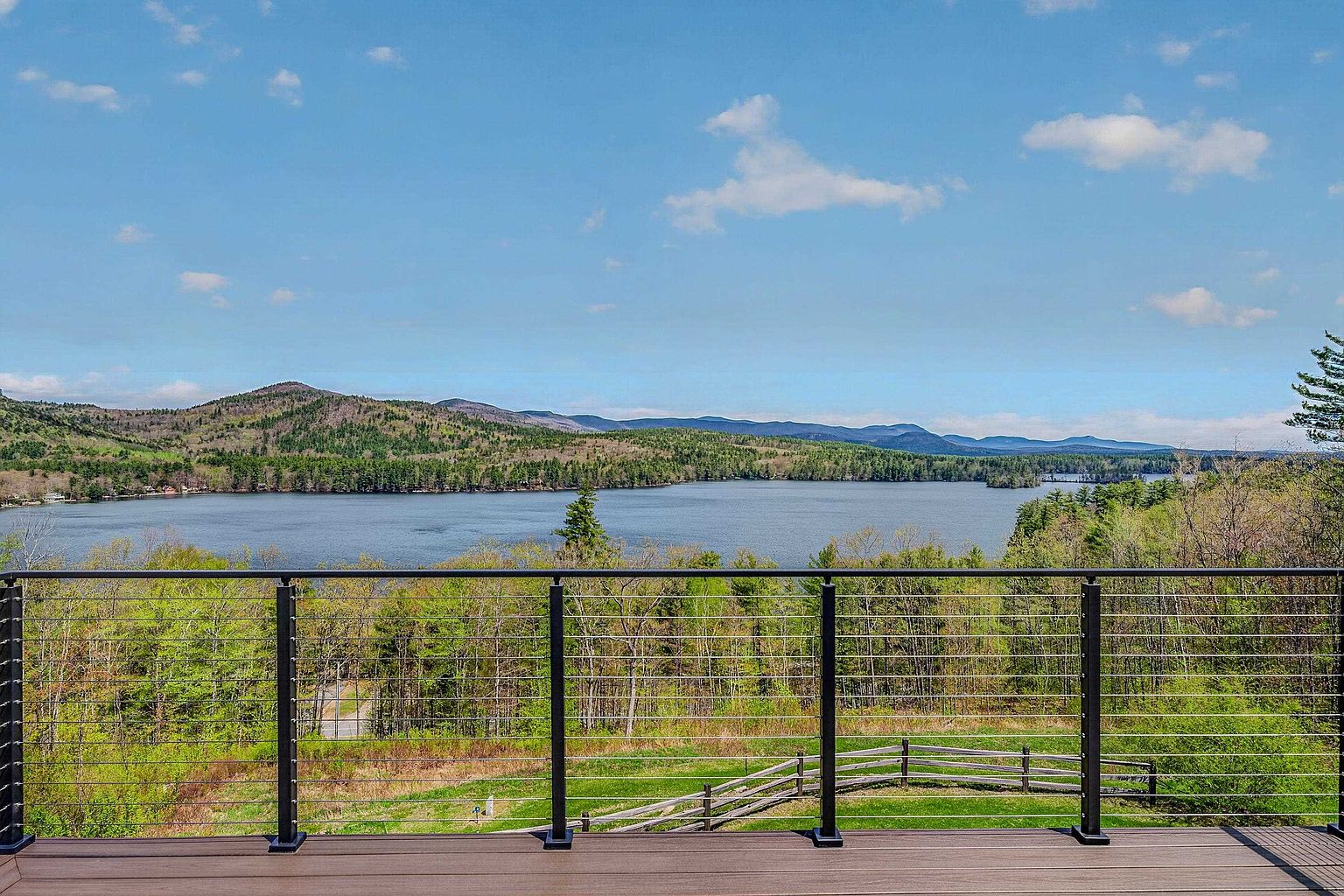This image captures a wide, elevated deck featuring modern composite decking and a sleek black metal railing with horizontal cable infill. The perspective looks out over a lush, green landscape toward a serene lake nestled against a backdrop of rolling mountains under a bright blue sky. The scene conveys a peaceful, scenic outdoor living space ideal for relaxation and taking in the expansive natural views.