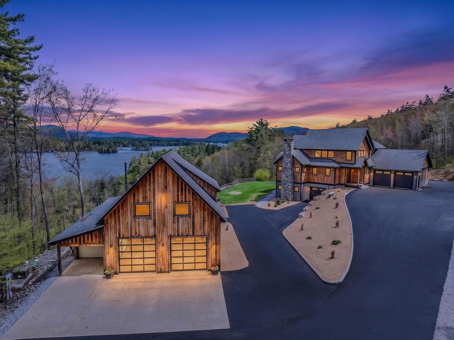 This elevated aerial perspective captures a stunning rustic-style estate at dusk, featuring a primary residence and a detached garage building set against a vibrant sunset over a lake. The property showcases natural wood siding, stone accents, and expansive paved driveways that wind through the landscaped grounds. The composition highlights the serene waterfront location and the architectural harmony between the two structures.