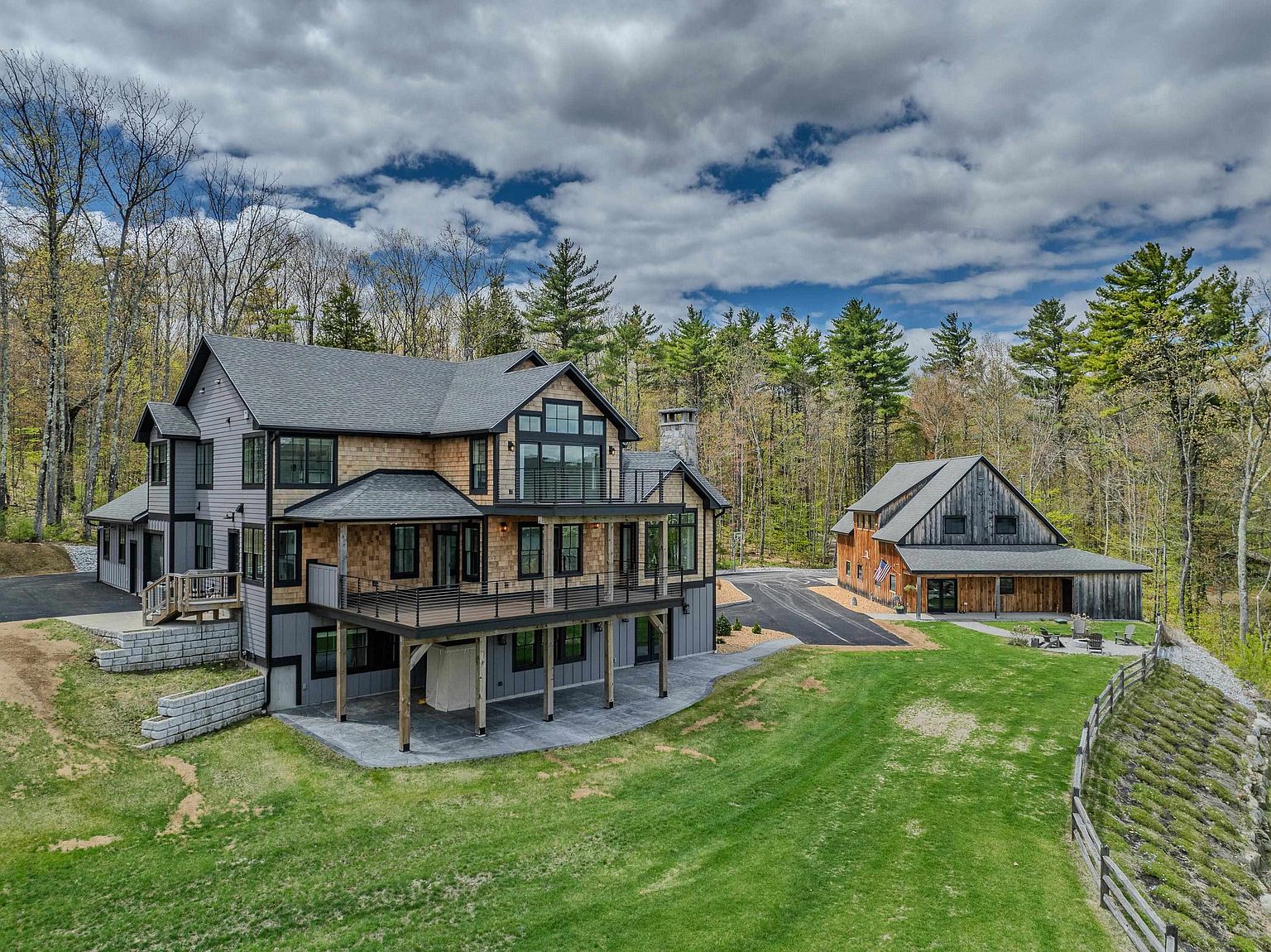 This aerial perspective captures a sprawling property featuring a large, multi-level custom home with a blend of gray siding and natural wood shingles. The residence includes an expansive elevated deck and a lower-level patio, while a separate, rustic-style barn structure sits nearby on the manicured lawn. Surrounded by a dense forest, the home presents a sophisticated mountain-modern aesthetic with clean lines and large windows.