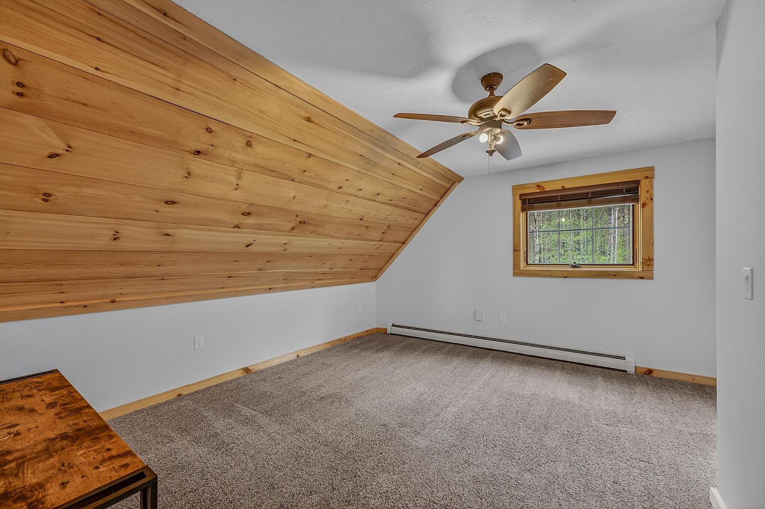 This bedroom features a unique slanted ceiling paneled in light wood, creating a cozy, rustic atmosphere. The room is carpeted and includes a window with wood trim that looks out onto a wooded area, along with a ceiling fan for climate control. A portion of a desk is visible in the foreground, suggesting the space could function as a bedroom or a home office.