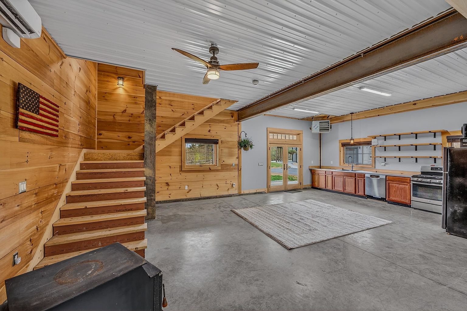This spacious, open-concept living area features a rustic aesthetic with light wood-paneled walls, a corrugated metal ceiling, and polished concrete floors. The room includes a wooden staircase, a kitchen area with wooden cabinetry and stainless steel appliances, and a large area rug centered in the space. A wood-burning stove sits in the foreground, and double glass doors provide natural light and access to the outdoors.