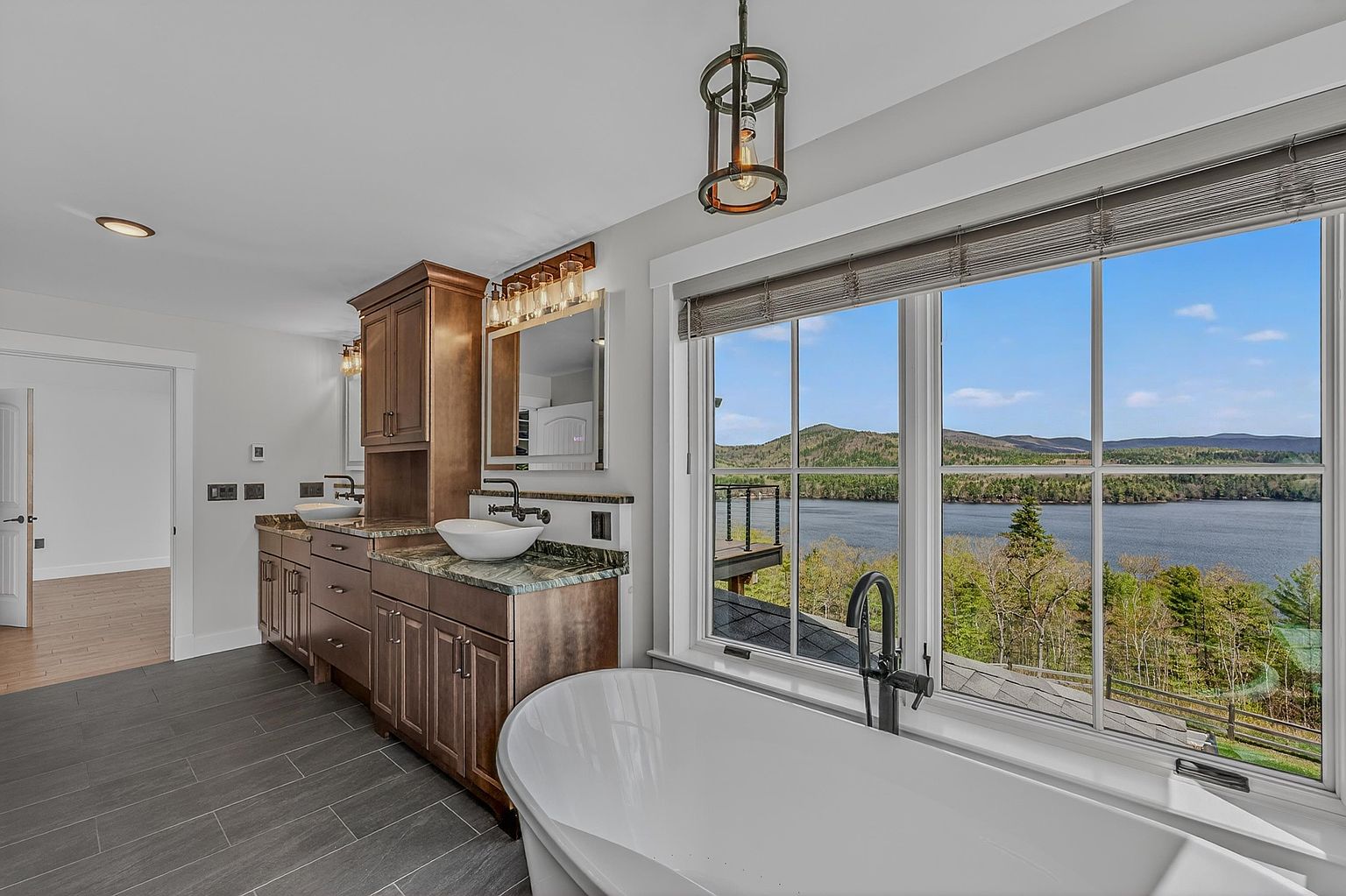 This primary bathroom features a luxurious freestanding soaking tub positioned directly in front of a large window that offers expansive views of a lake and surrounding mountains. The vanity area includes dual vessel sinks set into a stone countertop with dark wood cabinetry and a decorative pendant light overhead. The space is finished with neutral gray floor tiles, creating a serene and high-end atmosphere.